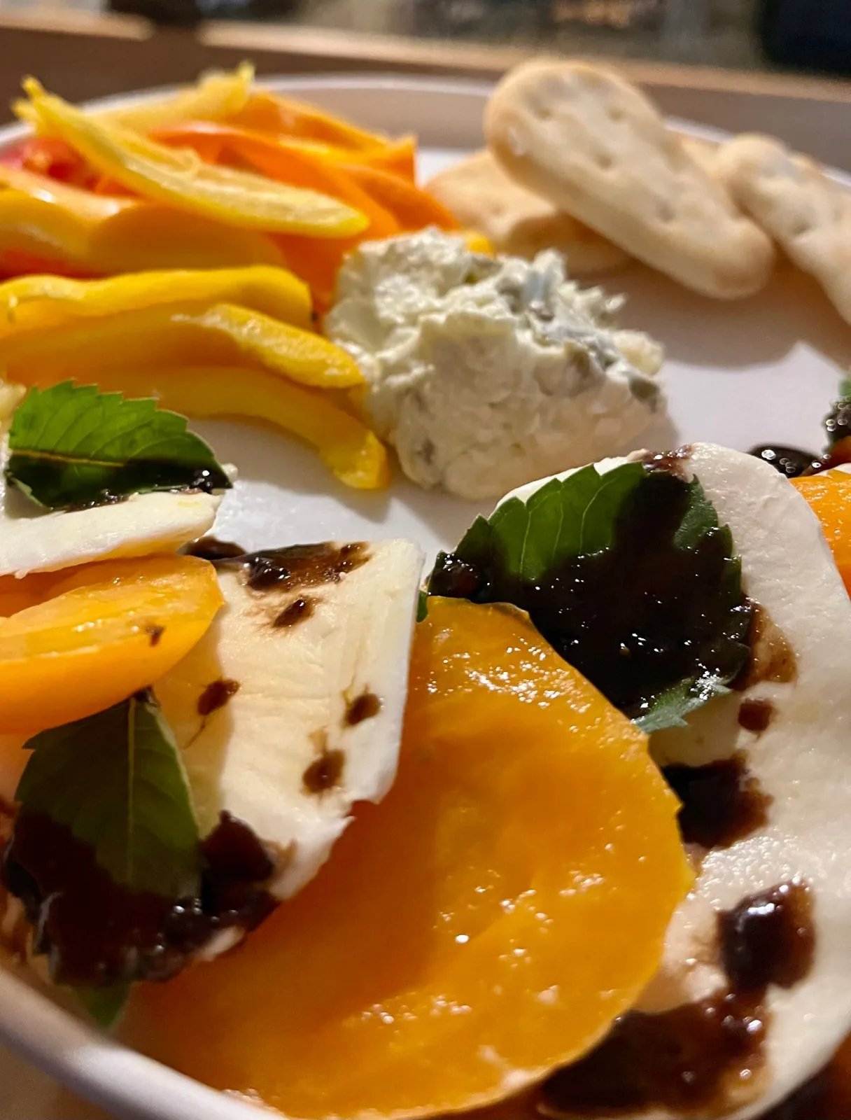 A plate of golden heirloom tomato caprese with fresh mozzarella, basil and balsamic, beside sliced sweet peppers and an herbed dip with crackers